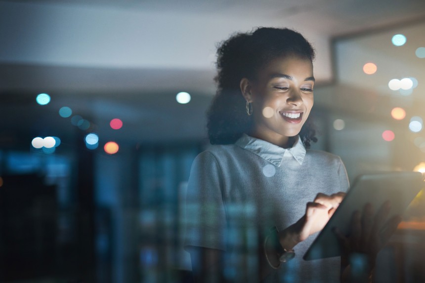 
			  	Junge Frau arbeitet l�chelnd nachts an einem Tablet, Lichtreflektionen und moderne B�roumgebung im Hintergrund.
			  