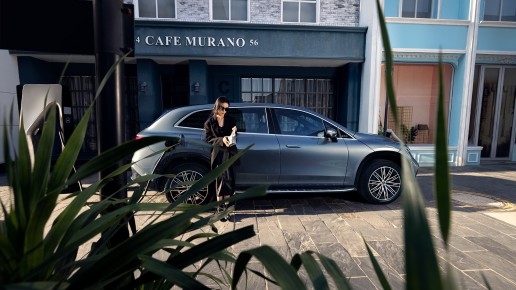 
						  Frau steht mit Kaffeebecher neben einem geparkten silbernen Mercedes-Benz EQ vor einem Caf�, im Vordergrund gr�ne Pflanzen.
						  