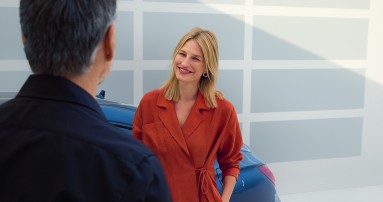 
						  Junge Frau in rotem Outfit steht l�chelnd vor einem Mann neben einem Mercedes-Benz Fahrzeug und f�hrt ein Gespr�ch.
						  