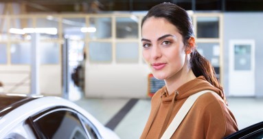 
						  Junge Frau in braunem Hoodie steht l�chelnd neben einem wei�en Mercedes-Benz in einer hellen Werkstattumgebung.
						  