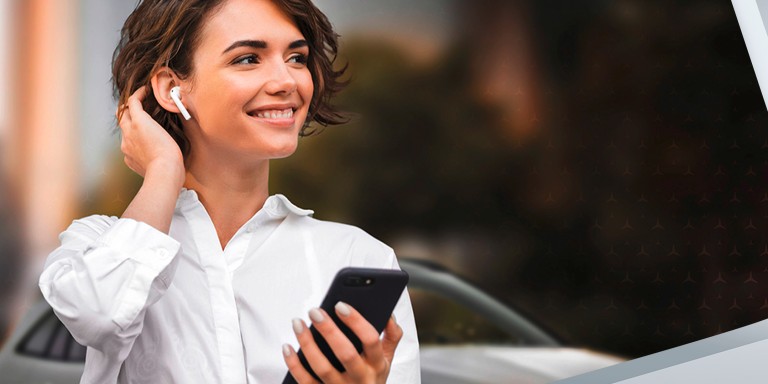 
							Junge Frau mit wei�en kabellosen Kopfh�rern h�lt ein Smartphone in der Hand und l�chelt, im Hintergrund ist ein Auto zu sehen.
					 	 