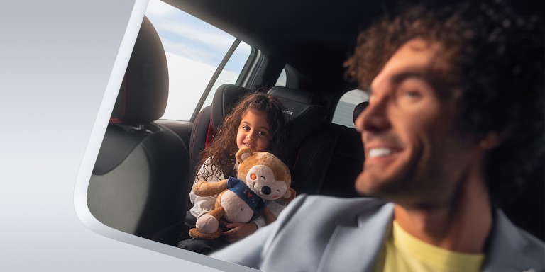 
							Mann sitzt lchelnd im Auto, whrend ein Kind im Kindersitz auf der Rckbank sitzt und ein Kuscheltier hlt.
					 	 