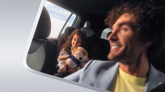 
							Mann sitzt l�chelnd im Auto, w�hrend ein Kind im Kindersitz auf der R�ckbank sitzt und ein Kuscheltier h�lt.
					 	 