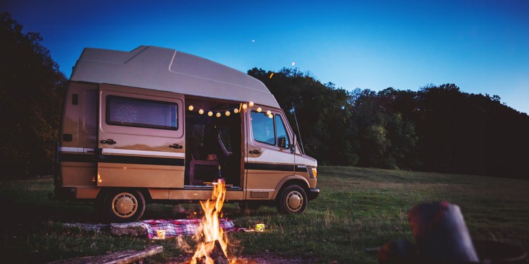 
				Ein Marco Polo, auf Basis des Mercedes-Benz 209 D, steht mit geffneter Seitentr auf einer Wiese im Abendlicht, vor dem Van brennt ein Lagerfeuer und aus dem Fahrzeuginneren hngt eine Lichterkette heraus.
				
