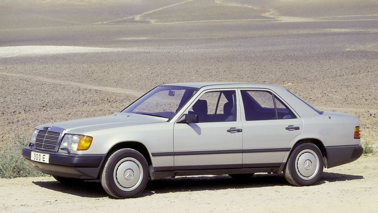 
			Ein Mercedes-Benz 300 E steht auf einer Schotterstrae, im Hintergrund karge Landschaft.
		