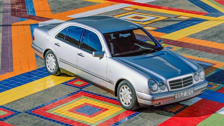 
			Ein Mercedes-Benz der Baureihe 210 parkt auf einem Platz mit bunten Fliesen.
		