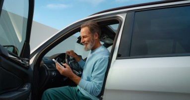 
						  Ein Mann sitzt bei geffneter Fahrertr am Steuer eines silbernen Mercedes-Benz Fahrzeugs und blickt lchelnd auf ein Smartphone in seiner Hand.
						  