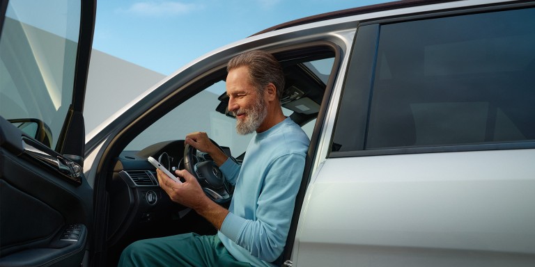 
				Ein Mann sitzt bei geffneter Fahrertr am Steuer eines silbernen Mercedes-Benz Fahrzeugs und blickt lchelnd auf ein Smartphone in seiner Hand.
				