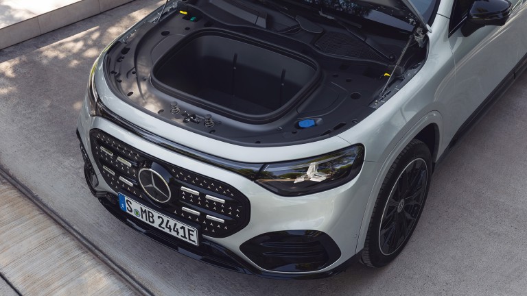 
			Das Bild zeigt die vollst�ndig ge�ffnete Frontklappe des GLB, unter der der zus�tzliche Stauraum im Frunk sichtbar ist. Das Fach ist mittig platziert und von schwarzen Kunststoffverkleidungen umgeben. Der Wagen steht im Freien.
		