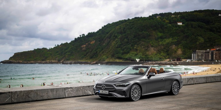 
				Ein Mercedes-Benz CLE Cabriolet in graphitgrau magno mit AMG-Line steht vor einem Badestrand an einer Meereskste.
				