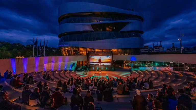
			Ein abendliches Konzert mit einem Publikum, das auf der Bhne vor dem Mercedes-Benz Museum Stuttgart einem Musiker lauscht.
		