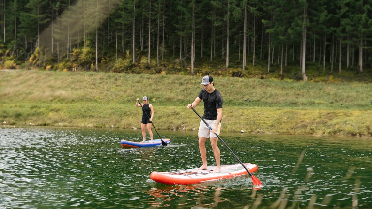 
			Ein Mann und eine Frau stehen auf SUP-Boards und paddeln ber einen See.
		