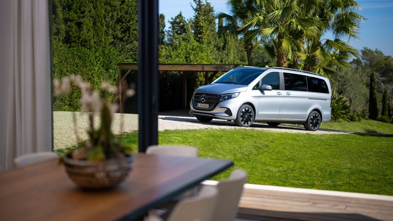 
			Blick vom Inneren eines Hauses auf eine Mercedes-Benz V-Klasse in hightechsilber metallic, die in der Einfahrt vor Palmen und einer hohen Hecke parkt.
		