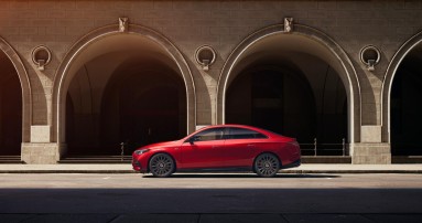 
						  Der neue Mercedes-Benz CLA mit EQ Technologie in Patagonienrot fhrt vor Arkaden auf einer Strae. 
						  