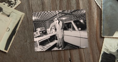 
						  Ein Schwarz-Wei-Bild aus 1970 liegt neben anderen alten Fotos auf einem Holztisch, auf dem Bild ist ein Mercedes-Benz Ingenieur zu sehen, der mit Kopfhrern in einem schallisolierten Raum neben einem alten Mercedes-Benz Fahrzeug steht und Akustiktests durchfhrt.
						  