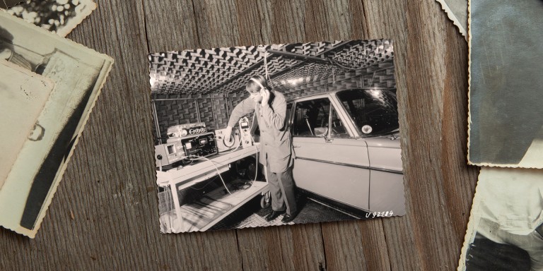 
				Ein Schwarz-Wei-Bild aus 1970 liegt neben anderen alten Fotos auf einem Holztisch, auf dem Bild ist ein Mercedes-Benz Ingenieur zu sehen, der mit Kopfhrern in einem schallisolierten Raum neben einem alten Mercedes-Benz Fahrzeug steht und Akustiktests durchfhrt.
				