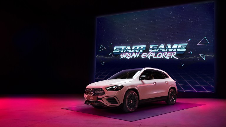 
			Ein weier GLA steht vor einer Leinwand, auf der der Schriftzug „Start Game Urban Explorer“ zu sehen ist.
		