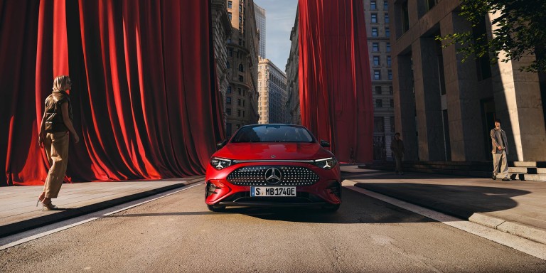 
				Ein roter CLA mit EQ Technologie steht auf einer Strae, im Hintergrund sind groe rote Vorhnge zu sehen, die von den Husern herabhngen. Links vom Fahrzeug steht eine Frau, rechts luft ein Mann auf dem Fugngerweg.
				