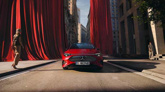 
				Ein roter CLA mit EQ Technologie steht auf einer Stra�e, im Hintergrund sind gro�e rote Vorh�nge zu sehen, die von den H�usern herabh�ngen. Links vom Fahrzeug steht eine Frau, rechts l�uft ein Mann auf dem Fu�g�ngerweg.
				