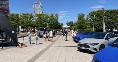 
						  Blick auf den Familientag im Werk Rastatt, links ist ein Pavillon zu sehen, rechts dahinter zwischen den Bumen ein Riesenrad, in der Mitte laufen Besuchende ber den Platz und rechts stehen vier CLA, einer in Rot, zwei in Blau und einer in Grau.
						  