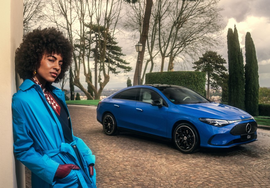 
			  	Eine Frau lehnt an einer hellen Wand, im Hintergrund steht ein Mercedes-Benz CLA in Klarblau auf einer gepflasterten Auffahrt, umgeben von einer Parklandschaft.
			  