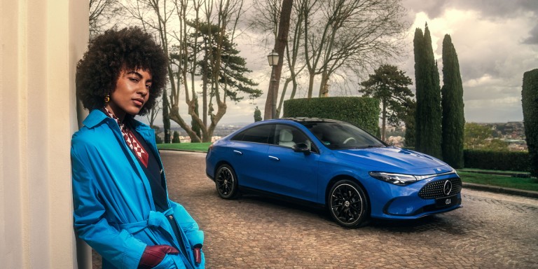 
							Eine Frau lehnt an einer hellen Wand, im Hintergrund steht ein Mercedes-Benz CLA in Klarblau auf einer gepflasterten Auffahrt, umgeben von einer Parklandschaft.
					 	 