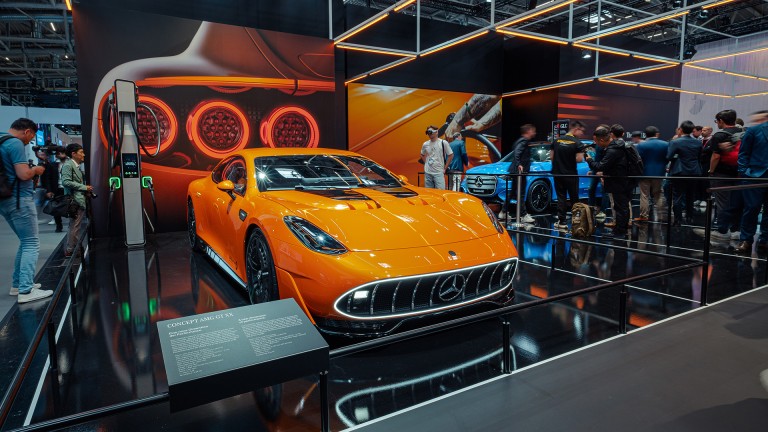 
			Orangefarbenes Concept Car Mercedes-AMG GT XX mittig auf einem Podest. Links und im Hintergrund stehen Besucherinnen und Besucher in der Messehalle.
		