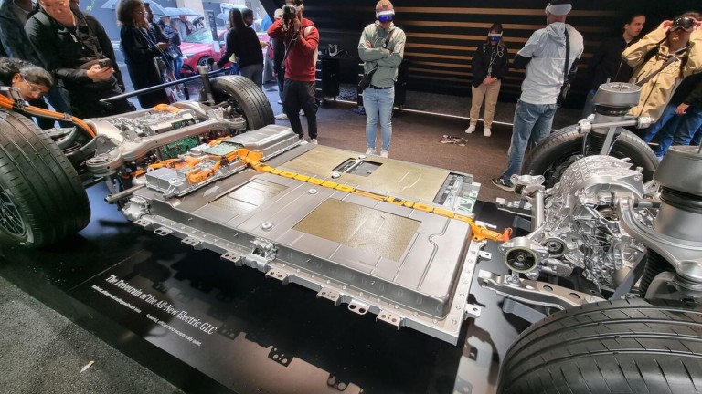
			Im Vordergrund ist ein elektrischer Antriebsstrang des Mercedes-Benz GLC mit Batterie und Achsen zu sehen. Im Hintergrund stehen Menschen, einige tragen VR-Brillen.
		