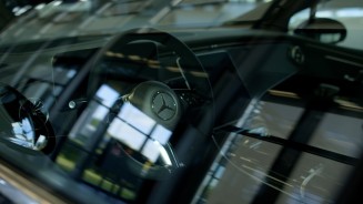 
Blick durch die Fensterscheibe in das Cockpit der Mercedes-Benz EQE Limousine mit dem Mercedes-Benz Lenkrad im Zentrum. Lichtspiegelungen auf der Fensterscheibe.
