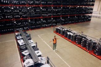 
Blick in die Wareneingangshalle: Im Hintergrund reihen sich meterhohe Regale voller R�der. Davor befinden zwei kleinere Regale, zwischen denen Ali Tas mit ausgebreiteten Armen steht. Er tr�gt eine Warnweste.
