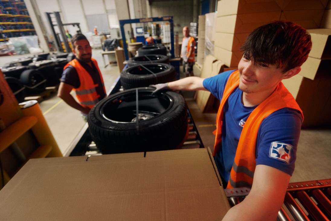 
			Am F�rderband werden R�der in Kartons verpackt, im Vordergrund l�chelt ein Mitarbeiter, w�hrend er das Rad in den Karton schiebt. Dahinter steht Ali Tas. Beide tragen Warnwesten.
		