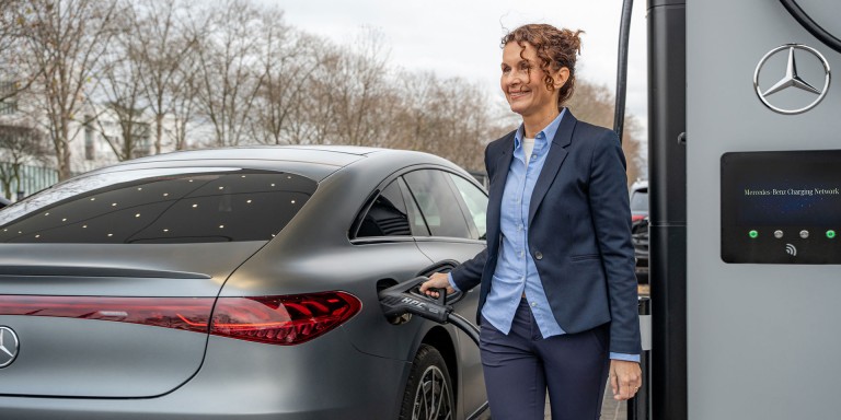 
				Eine Frau in dunkelblauer Office-Kleidung ldt ihren vollelektrischen Mercedes-Benz an der Ladesule eines Mercedes-Benz Charging Hubs auf.
				