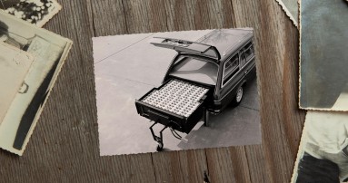 
						  Ein Schwarz-Wei-Bild von 1982 liegt neben anderen alten Fotos auf einem Holztisch. Auf dem Bild ist der geffnete Kofferraum eines Mercedes-Benz T-Modells der Baureihe 123 zu sehen. Im Kofferraum ist eine groe Nickel-Eisen-Batterie sichtbar, die fast den gesamten Platz einnimmt und herausgezogen auf einem Sttzrad steht. Bildquelle: Mercedes-Benz Classic.
						  