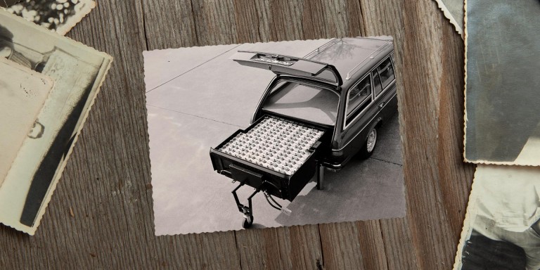 
				Ein Schwarz-Wei-Bild von 1982 liegt neben anderen alten Fotos auf einem Holztisch. Auf dem Bild ist der geffnete Kofferraum eines Mercedes-Benz T-Modells der Baureihe 123 zu sehen. Im Kofferraum ist eine groe Nickel-Eisen-Batterie sichtbar, die fast den gesamten Platz einnimmt und herausgezogen auf einem Sttzrad steht. Bildquelle: Mercedes-Benz Classic.
				