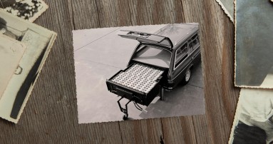 
					  Ein Schwarz-Wei�-Bild von1982 liegt neben anderen alten Fotos auf einem Holztisch. Auf dem Bild ist der ge�ffnete Kofferraum eines Mercedes-Benz T-Modells der Baureihe 123 zu sehen. Im Kofferraum ist eine gro�e Nickel-Eisen-Batterie sichtbar, die fast den gesamten Platz einnimmt und herausgezogen auf einem St�tzrad steht. Bildquelle: Mercedes-Benz Classic.
					  