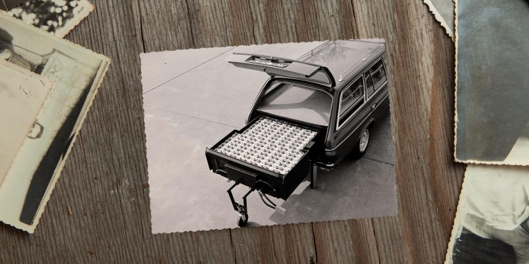 
				Ein Schwarz-Wei�-Bild von1982 liegt neben anderen alten Fotos auf einem Holztisch. Auf dem Bild ist der ge�ffnete Kofferraum eines Mercedes-Benz T-Modells der Baureihe 123 zu sehen. Im Kofferraum ist eine gro�e Nickel-Eisen-Batterie sichtbar, die fast den gesamten Platz einnimmt und herausgezogen auf einem St�tzrad steht. Bildquelle: Mercedes-Benz Classic.
				