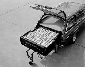 
Schwarz-Wei-Aufnahme des geffneten Kofferraums eines Mercedes-Benz T-Modells der Baureihe 123. Im Kofferraum ist die groe Nickel-Eisen-Batterie sichtbar, die fast den gesamten Platz einnimmt und herausgezogen auf einem Sttzrad steht.
