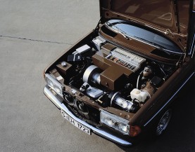 
Farbaufnahme des geffneten Motorraums eines Mercedes-Benz T-Modells der Baureihe 123. In dem Versuchsfahrzeug sind statt klassischem Motorblock Kabel, Aggregate und ein groformatiger Batterieblock sichtbar
