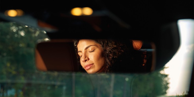 
				Nahaufnahme eines Rckspiegels, in dem eine Frau auf dem Rcksitz eines Mercedes-Benz zu sehen ist, wie sie mit geschlossenen Augen die Sonne geniet.
				