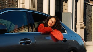 
Eine braunhaarige Frau mit rotem Pullover sitzt auf dem Rcksitz eines Mercedes-Benz Fahrzeugs, lehnt sich mit geschlossenen Augen leicht aus dem Fenster und hat die verschrnkten Arme auf der Fensterffnung abgelegt.
