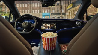 
Blick vom Rcksitz auf die Mittelkonsole eines Mercedes-Benz Fahrzeugs. Auf dem Zentraldisplay luft ein Film, und auf der Mittelablage steht ein mit Popcorn gefllter rot-wei-gestreifter Pappeimer. Auf dem Beifahrersitz ist der linke Rand einer Pizza zu sehen.
