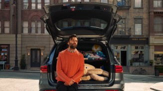 
Ein Mann mit einem orangeroten Pullover sitzt auf dem Rand des geffneten und voll beladenen Kofferraums eines Mercedes-Benz Fahrzeugs und hat die Augen geschlossen. Hinter dem Fahrzeug ist eine urbane Huserfassade mit Lden zu sehen.
