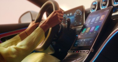 
					  Die Hnde einer Frau umfassen das Lenkrad eines Mercedes-Benz, im Vordergrund ist das Zentraldisplay mit MBUX zu sehen. Auf dem Bild ist in groen, weien Buchstaben der Schriftzug „Hey Mercedes!“ zu lesen.
					  