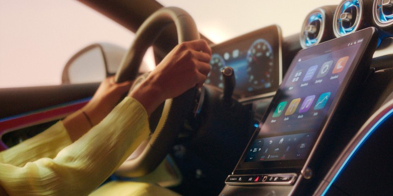 
				Die H�nde einer Frau umfassen das Lenkrad eines Mercedes-Benz, im Vordergrund ist das Zentraldisplay mit MBUX zu sehen. Auf dem Bild ist in gro�en, wei�en Buchstaben der Schriftzug &bdquo;Hey Mercedes!&ldquo; zu lesen.
				