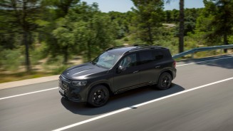 
Ein Mercedes-Benz GLB in MANUFAKTUR kosmosschwarz magno f�hrt auf einer kurvigen Stra�e durch eine gr�ne, bewaldete Landschaft.
