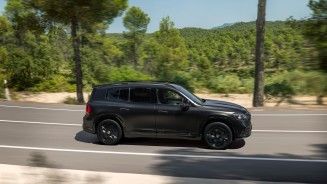 
Der elektrische Mercedes-Benz GLB in MANUFAKTUR kosmosschwarz magno f�hrt auf einer Stra�e durch eine bewaldete Landschaft.
