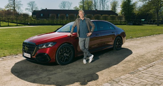 
					  Matthias Malmedie steht l�chelnd an der Fahrerseite neben der neuen S-Klasse von Mercedes-Benz in MANUFAKTUR patagonienrot metallic, die auf einem Weg inmitten einer gr�nen Wiese und B�umen steht.
					  