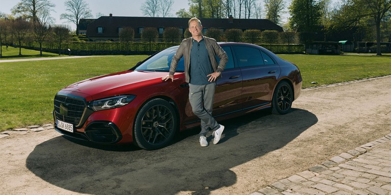 
				Matthias Malmedie steht l�chelnd an der Fahrerseite neben der neuen S-Klasse von Mercedes-Benz in MANUFAKTUR patagonienrot metallic, die auf einem Weg inmitten einer gr�nen Wiese und B�umen steht.
				
