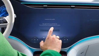 
Innenansicht eines modernen Fahrzeugs mit gro�em zentralen Touchscreen. Auf dem Bildschirm wird ein automatisches Software-Update angek�ndigt, w�hrend eine Person vom Fahrersitz aus mit dem Finger die Best�tigungstaste ber�hrt. Das Fahrzeug ist w�hrend des Updates laut Anzeige nicht nutzbar.
