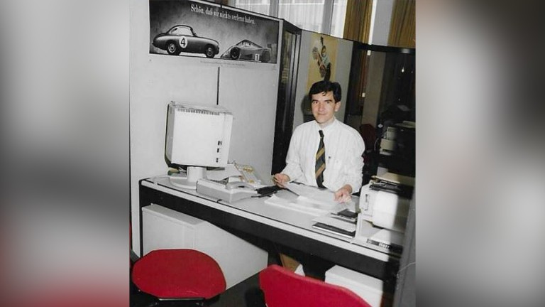 
			Ein Bild aus 1989 zeigt den ehemaligen Mercedes-Benz Members Mitarbeiter Eckhard Dunkel an seinem damaligen Beraterplatz. Rechts neben ihm steht ein alter Computer, vor seinem Tisch stehen zwei rote St�hle und er sitzt hinter dem Tisch und grinst in die Kamera.
		